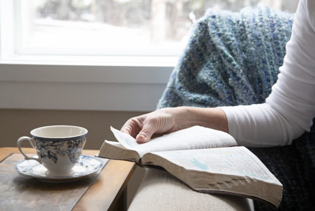 a woman sitting by a window reading a Bible with a cup of coffee nearby.